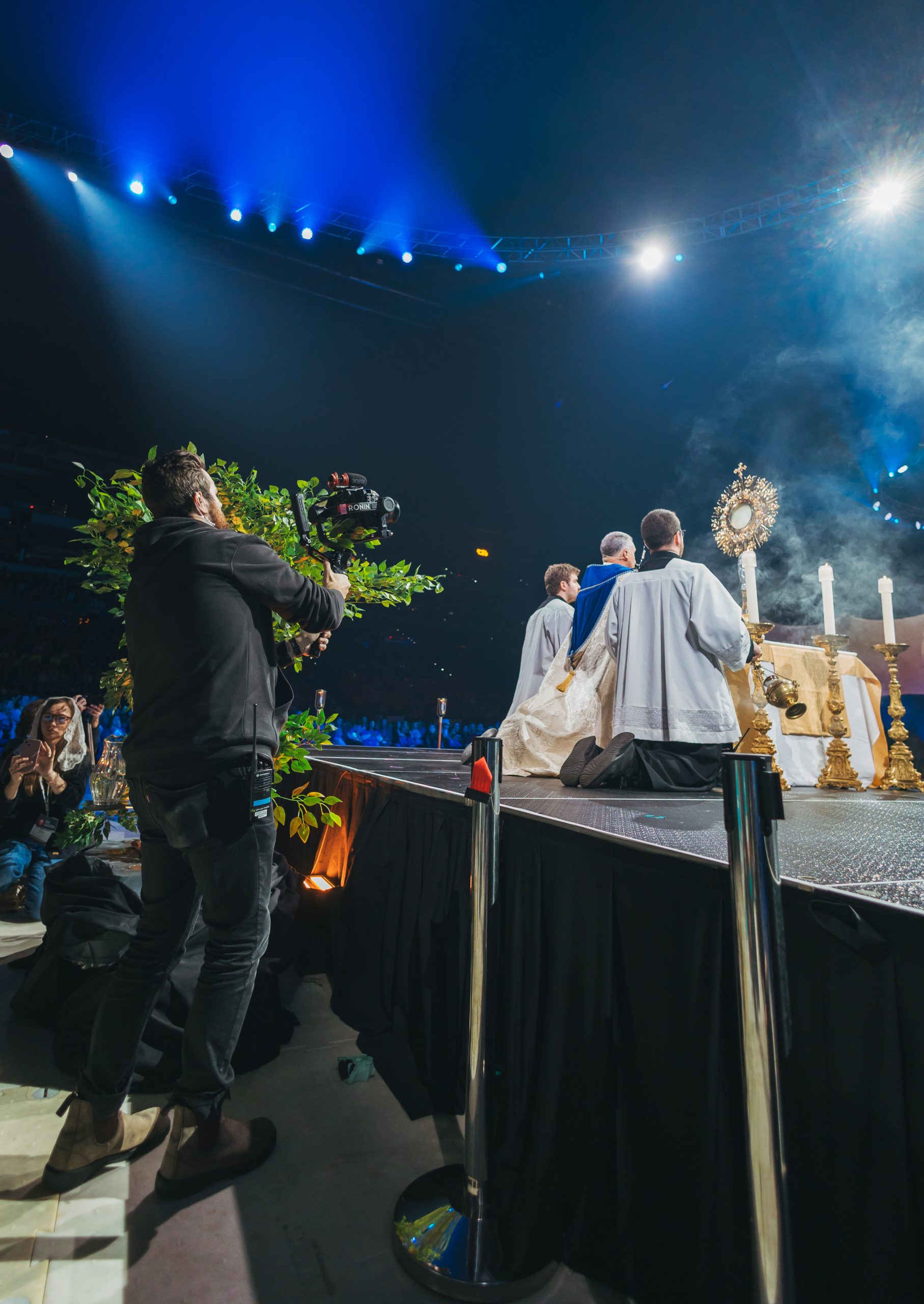 A filmmaker captures priests kneeling in Eucharistic adoration on a stage using a stabilized camera rig, showcasing reverent catholic video production during a live worship event.
