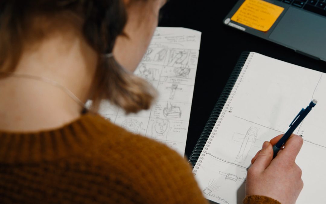 A creative team member sketches a storyboard beside production notes and a laptop, illustrating script and storyboard best practices for thoughtful Catholic video production planning.