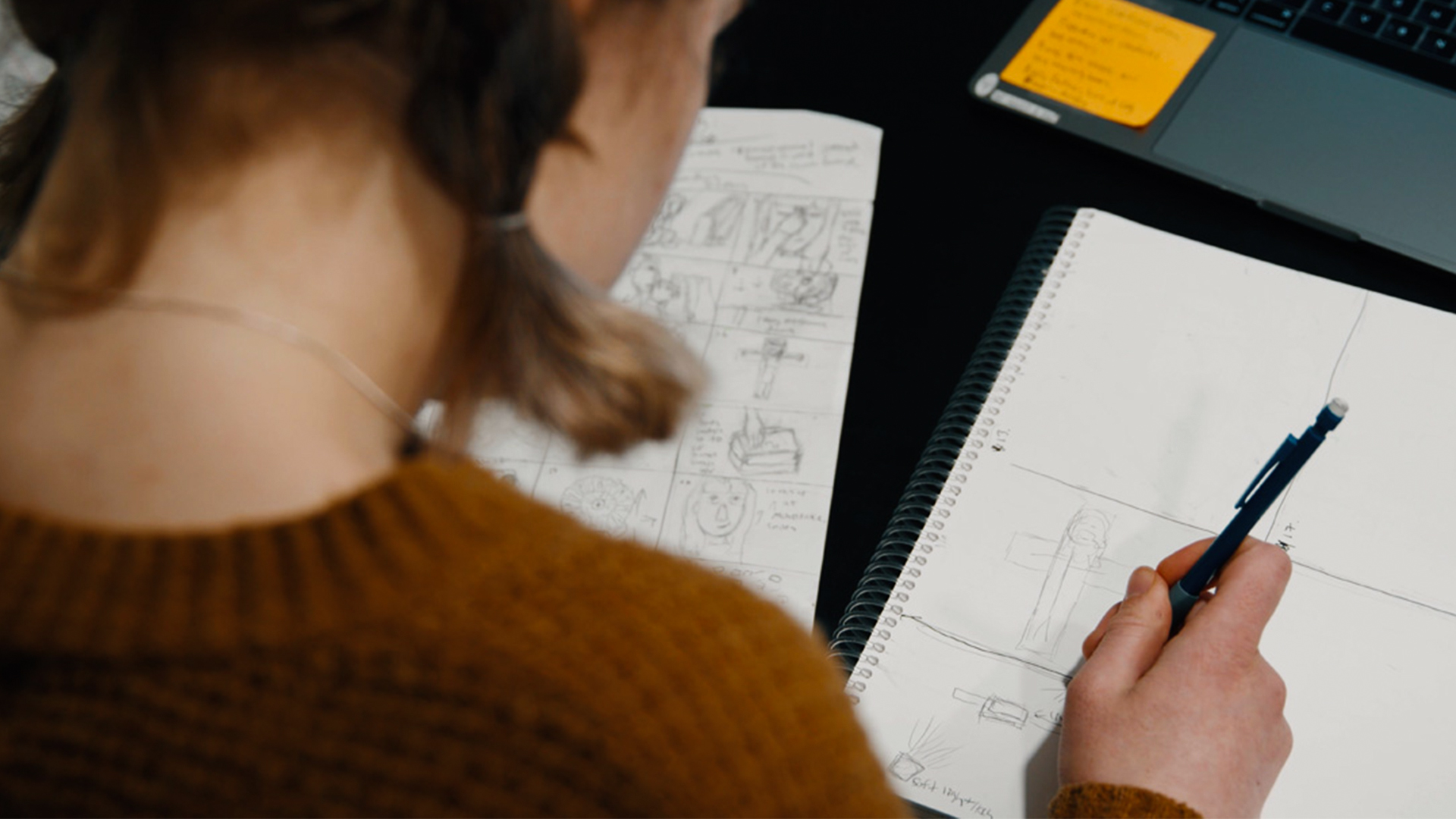 A creative team member sketches a storyboard beside production notes and a laptop, illustrating script and storyboard best practices for thoughtful Catholic video production planning.